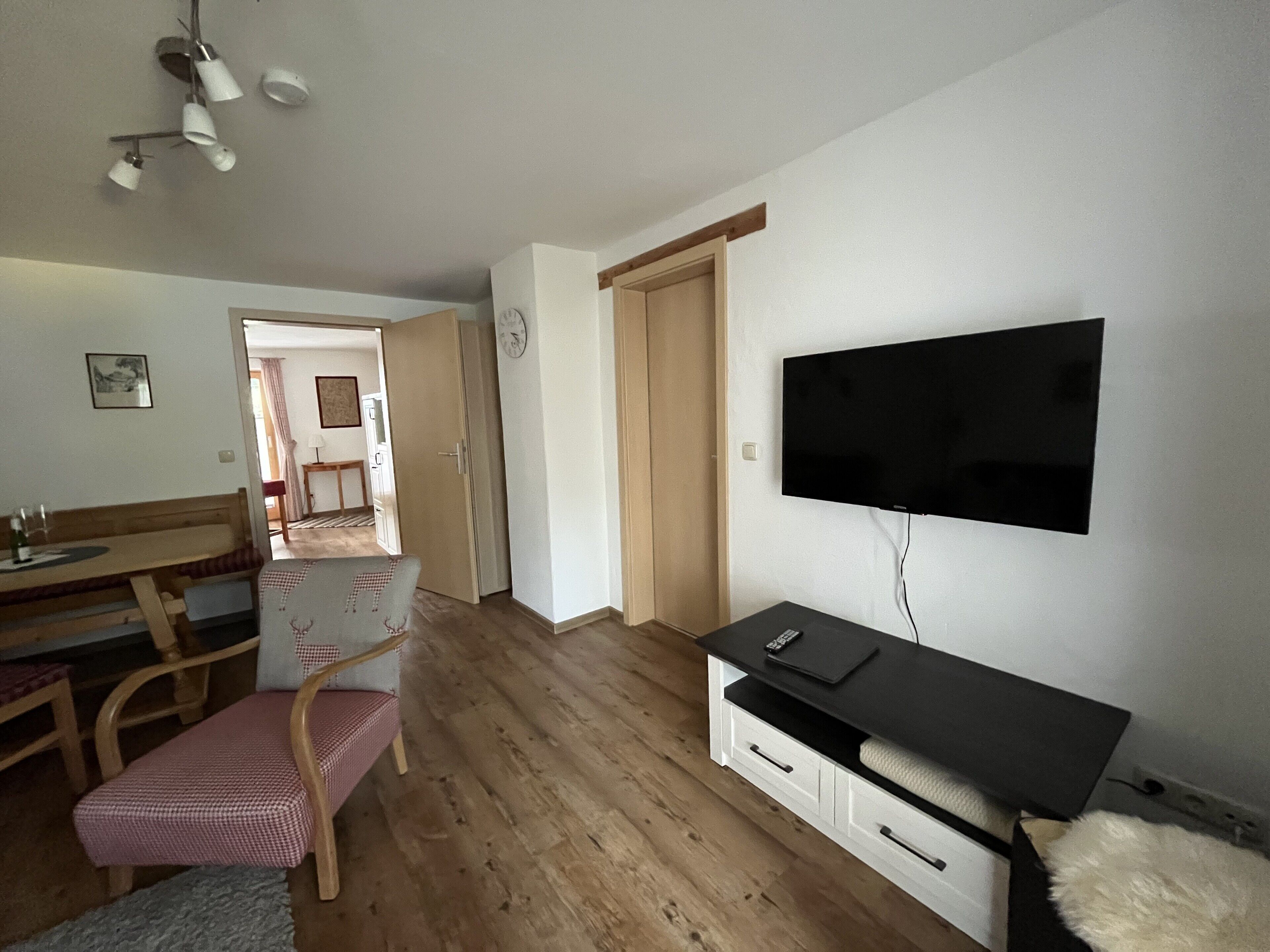 Una habitación con una televisión de pantalla plana montada en la pared, una mesa de comedor de madera con sillas, un pequeño sofá y suelo de madera.