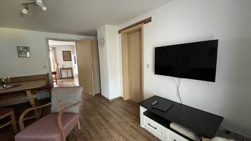 Una habitación con una televisión de pantalla plana montada en la pared, una mesa de comedor de madera con sillas, un pequeño sofá y suelo de madera.