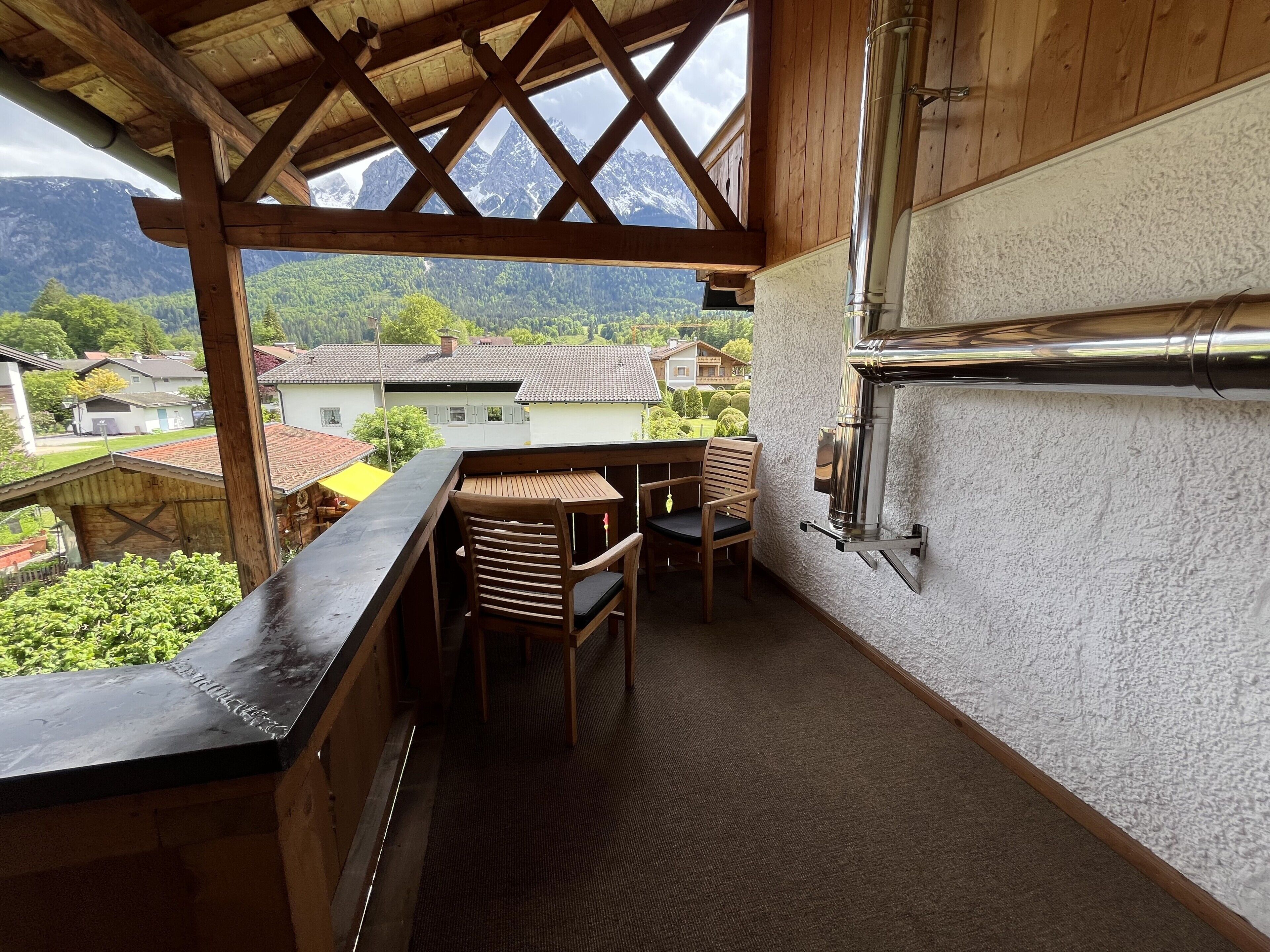 Un balcone in legno con vista su montagne e case, un tavolo di legno con sedie e un fornello in metallo.
