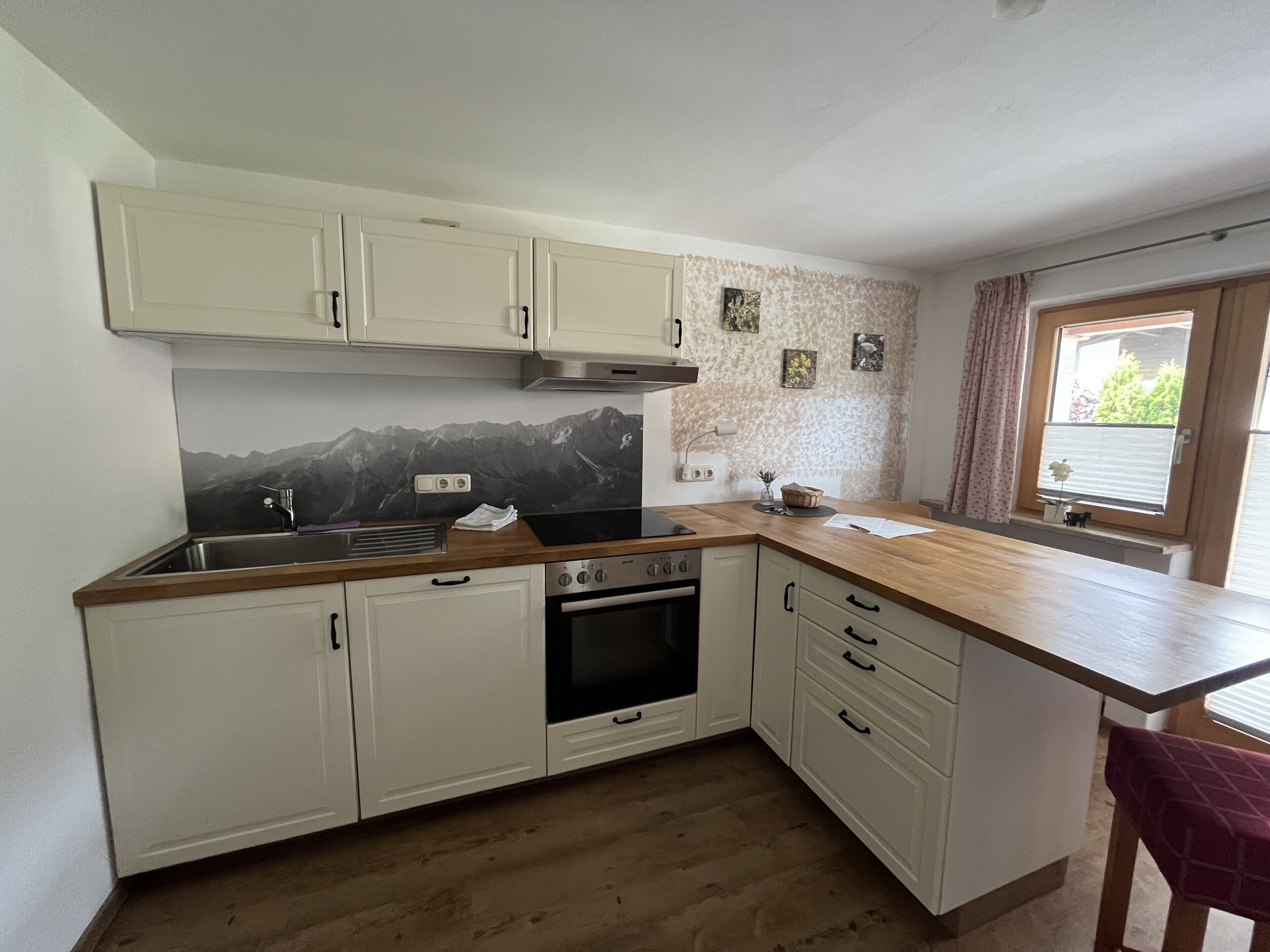 Une cuisine avec des meubles blancs, un plan de travail en bois, une cuisinière et une fenêtre donnant sur un paysage verdoyant.