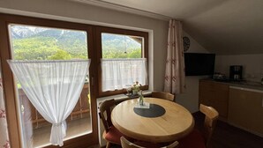 Ein Essbereich mit einem runden Holztisch, Stühlen, einem kleinen Topfpflanzen und Blick auf eine gebirgige Landschaft durch große Fenster mit transparenten Vorhängen.