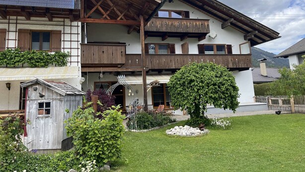 Eine zweistöckige Chalet mit Holzbalkonen, ein Garten mit Baum und Blumen, und eine kleine Holzkonstruktion.