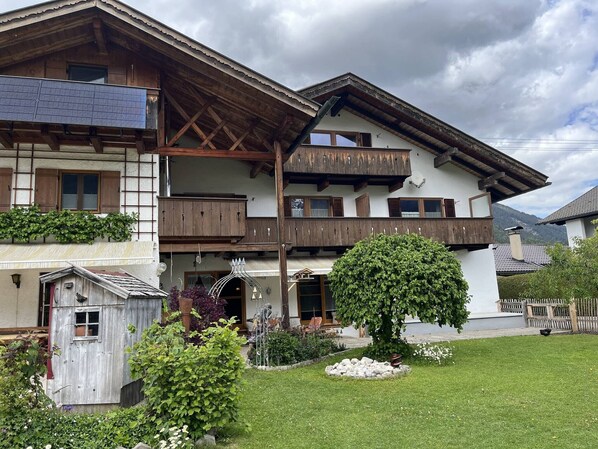 A two-story chalet with wooden balconies, a garden with a tree and flowers, and a small wooden structure. - Apartment 2 with Mountain View, Balcony, and Wi-Fi (Grainau)