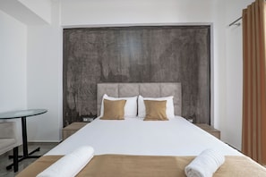 A hotel room with a large bed, two pillows, a bedside table, a small table, and a curtain.
