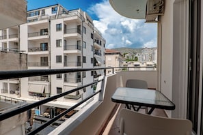 A balcony with a table and chairs, overlooking a multi-story building with balconies and a mountainous backdrop. - Grace Corner (Sarande)
