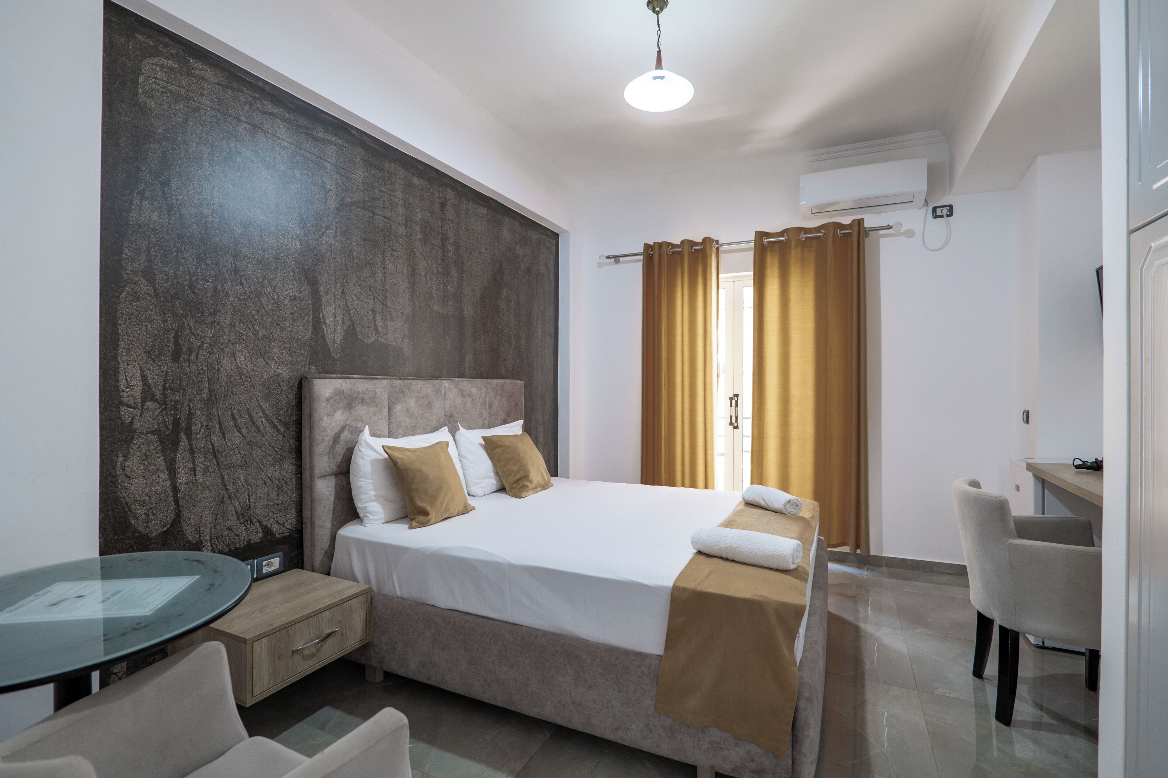 Camera d'albergo con un letto, un tavolino in vetro, una sedia, una scrivania e una finestra con tende.
