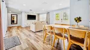 Uma sala de estar moderna com um sofá, uma mesa de jantar com cadeiras, uma televisão de tela plana na parede e uma janela com cortinas.