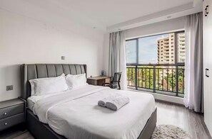 A modern hotel room with a large bed, a desk, and a balcony with a view of buildings. - Spacious Fully-stocked modern studio with a gym, cafe, & pitch in Kileleshwa (Nairobi)