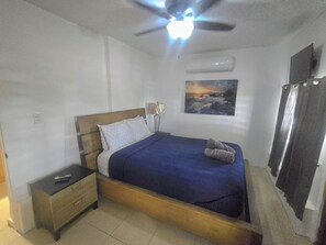 Un dormitorio con cama, mesita de noche, lámpara, equipo de aire acondicionado y un cuadro en la pared.