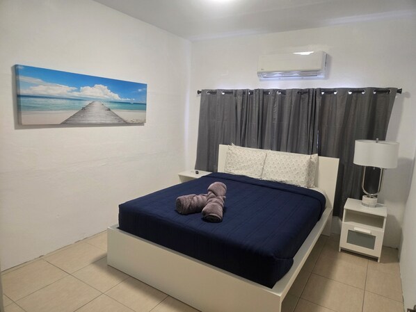 Un dormitorio con cama, un cuadro de playa, mesita de noche, lámpara y un equipo de aire acondicionado.