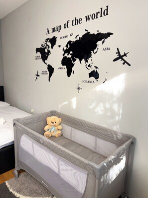 A baby crib with a teddy bear inside, a world map wall decal, and a bed with a white mattress. - Chopin Residence 6 - Cozy stay near Skopje center (Skopje)