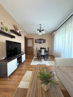 A modern living room with a flat-screen TV, wooden flooring, a dining area with a table and chairs, a ceiling fan, and a large window with curtains. - Chopin Residence 6 - Cozy stay near Skopje center (Skopje)