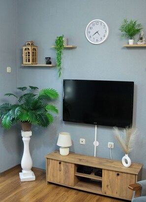 Un salon avec un téléviseur à écran plat fixé au mur, des étagères en bois ornées d’objets décoratifs, une plante en pot, un vase blanc et un meuble en bois.