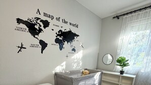 Ein Schlafzimmer mit Wanddeko in Form einer Weltkarte, einem Bett mit weißer Tagesdecke, einem grauen Stauraum, einem Nachttisch mit Spiegel und einem Fenster mit durchsichtigen Vorhängen.