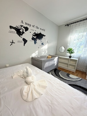 Een slaapkamer met een wereldkaart muurstickers, een bed met een witte dekbedovertrek, een grijze opbergunit, een nachtkastje met een spiegel, en een raam met dunne gordijnen.