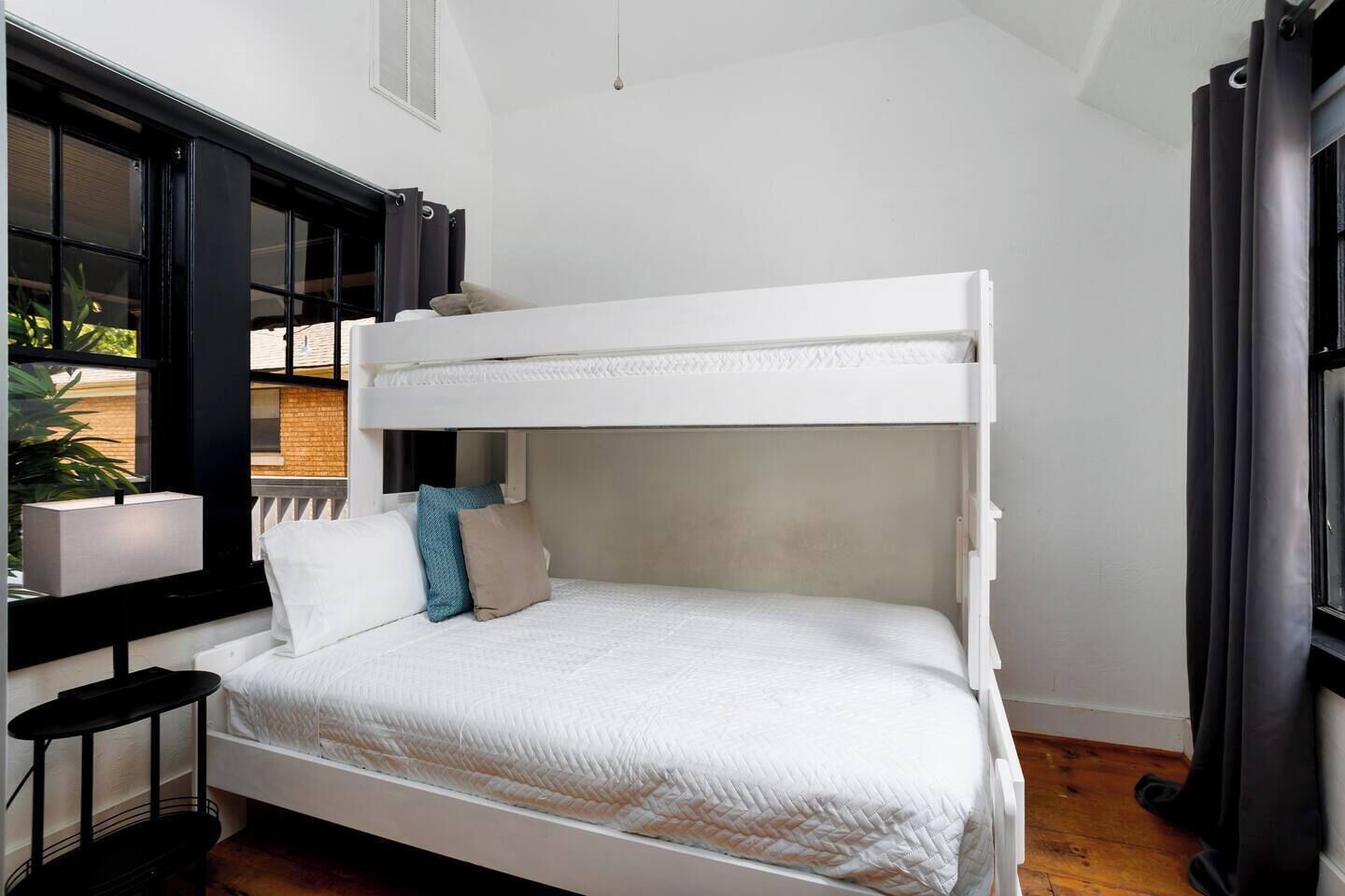 A bunk bed with white bedding, a bedside table, a lamp, and a window with curtains.