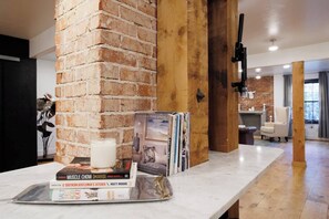 Ein modernes Wohnzimmer mit freiliegenden Ziegeln, einer Holzsäule, einem weißen Marmortisch mit Büchern und einer Kerze sowie einem Kamin im Hintergrund.