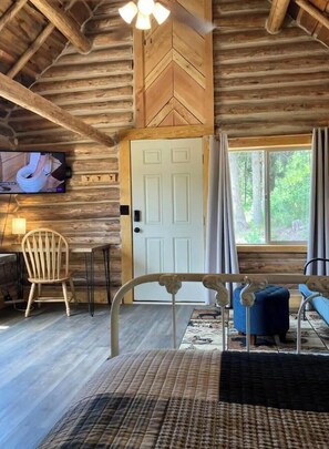 Ein Blockhausszimmer mit einem Bett, einem Sessel, einem Tisch, einem Fernseher und einem Fenster mit Vorhängen.
