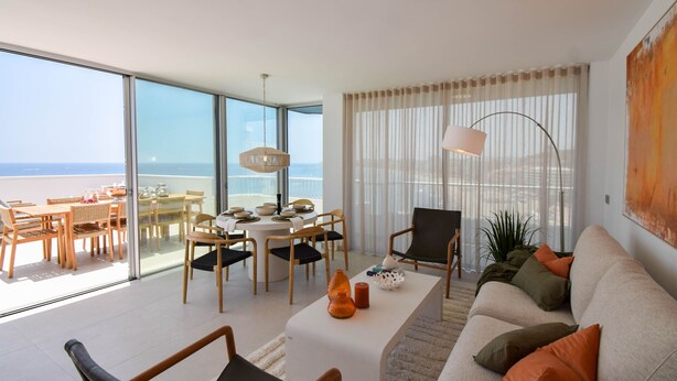 Ein modernes Wohnzimmer mit einem großen Glasfenster zum Balkon, einem runden Esstisch und einer Couch mit Kissen.