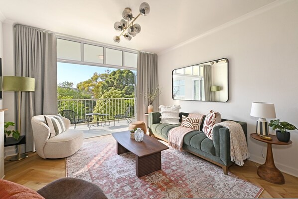 A spacious living room with a large window, a sofa, a coffee table, and a chandelier. - Two Bed Green Point Secure Designer Apartment (Cape Town)