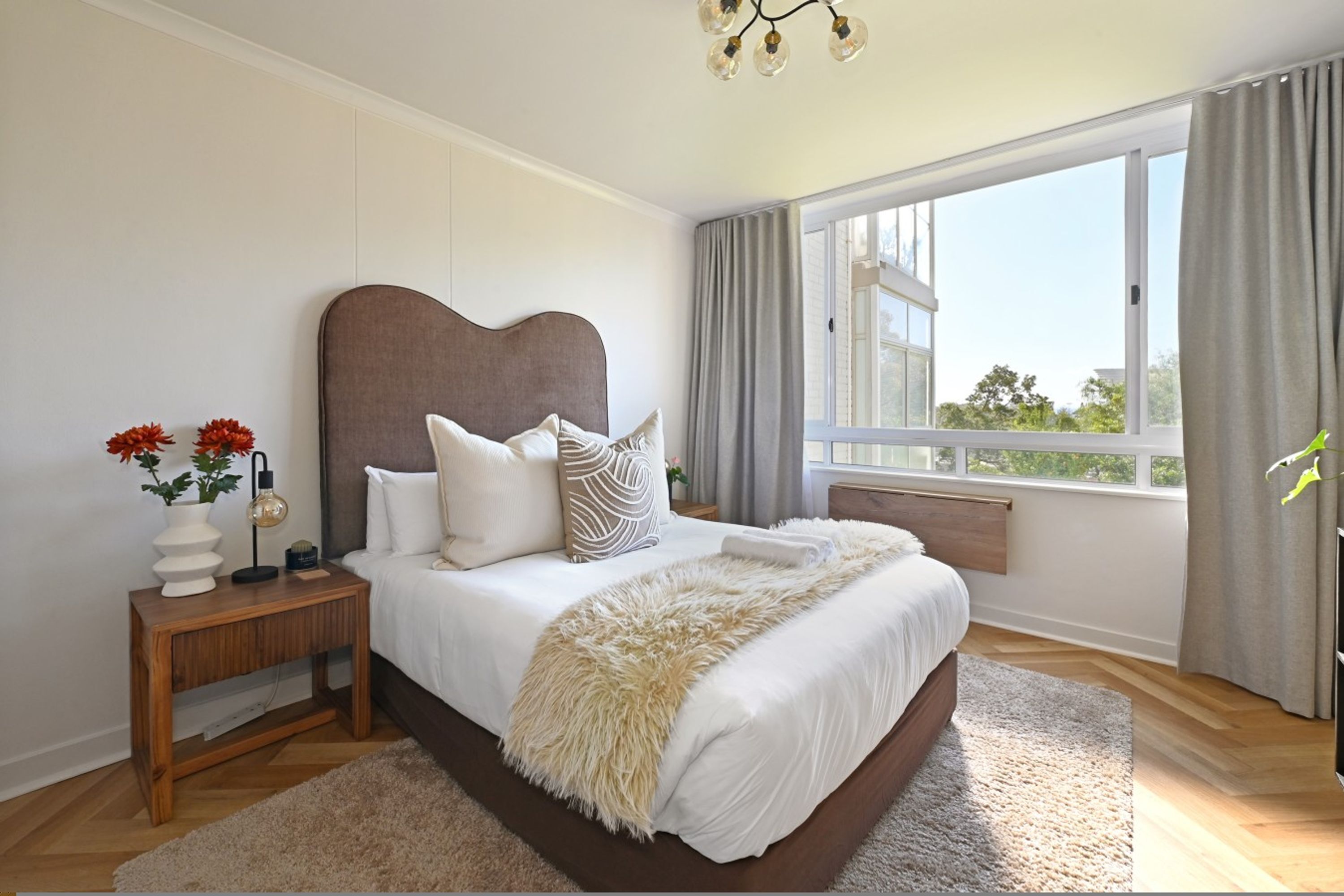 A bedroom with a large bed, a nightstand, a lamp, a vase with flowers, and a window with curtains.