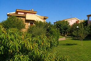 A building with a stone facade, a tiled roof, and a well-maintained garden with green shrubs and a lawn. - Vrbo Property (Tanaunella, Budoni)