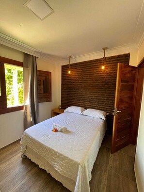 Ein Schlafzimmer mit einem Bett, einem Holzkopfende, einem Nachttisch, einem Fenster mit Vorhängen und einer Tür.