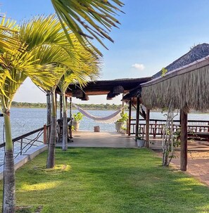 A wooden deck with a thatched roof, overlooking a body of water, surrounded by palm trees and potted plants. - Rustic house for family and friends, pets welcome. (Juritianha)