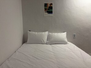 Habitació senzilla amb un llit, dos almoçons, un interruptor de llum fixat a la paret i un cartell a la paret.