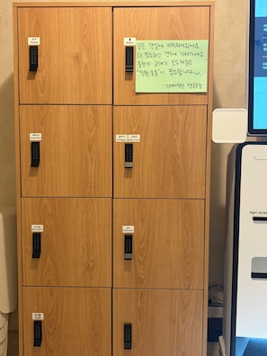 A wooden cabinet with multiple compartments, each labeled with a different amenity such as towel, water, charger, hair iron, board game, and toilet paper. - StayDucksan Hotel (Suwon)