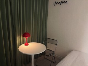 Una taula petita i rodona amb una làmpada vermella, una cadira de metall i un llit en una habitació amb cortines verdes.