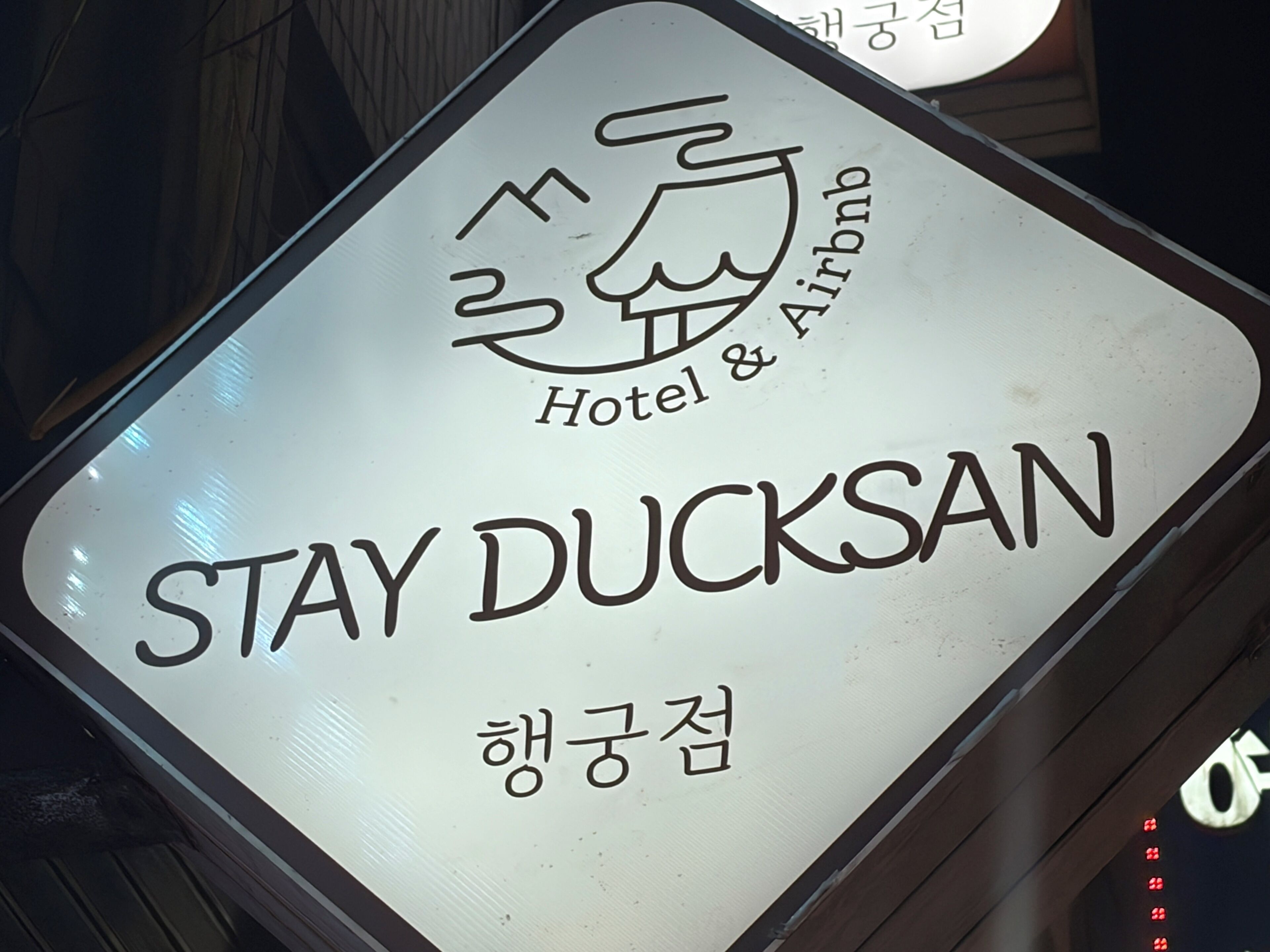 Een bord met 'Stay Ducksan Hotel & Airbnb', een logo en Koreaanse tekst.