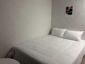 Un dormitori senzill i sense adorns, amb un llit individual, una petita taula de nit i un quadre penjat a la paret.