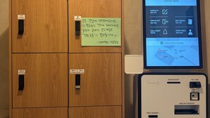 A self-check-in/out kiosk with a screen displaying instructions in Korean, a wooden cabinet with labeled compartments, and a sign with handwritten notes. - StayDucksan Hotel (Suwon)