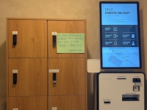 A self-check-in/out kiosk with a screen displaying instructions in Korean, a wooden cabinet with labeled compartments, and a sign with handwritten notes. - StayDucksan Hotel (Suwon)