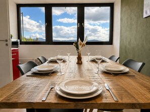 Une table à manger dressée pour six personnes, avec assiettes, verres et couverts, placée devant une fenêtre offrant une vue sur les montagnes.