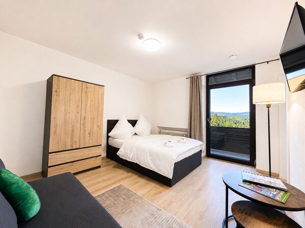 Une chambre moderne avec un lit, une armoire en bois, une petite table avec un magazine, et une vue sur la verdure grâce à une grande fenêtre.
