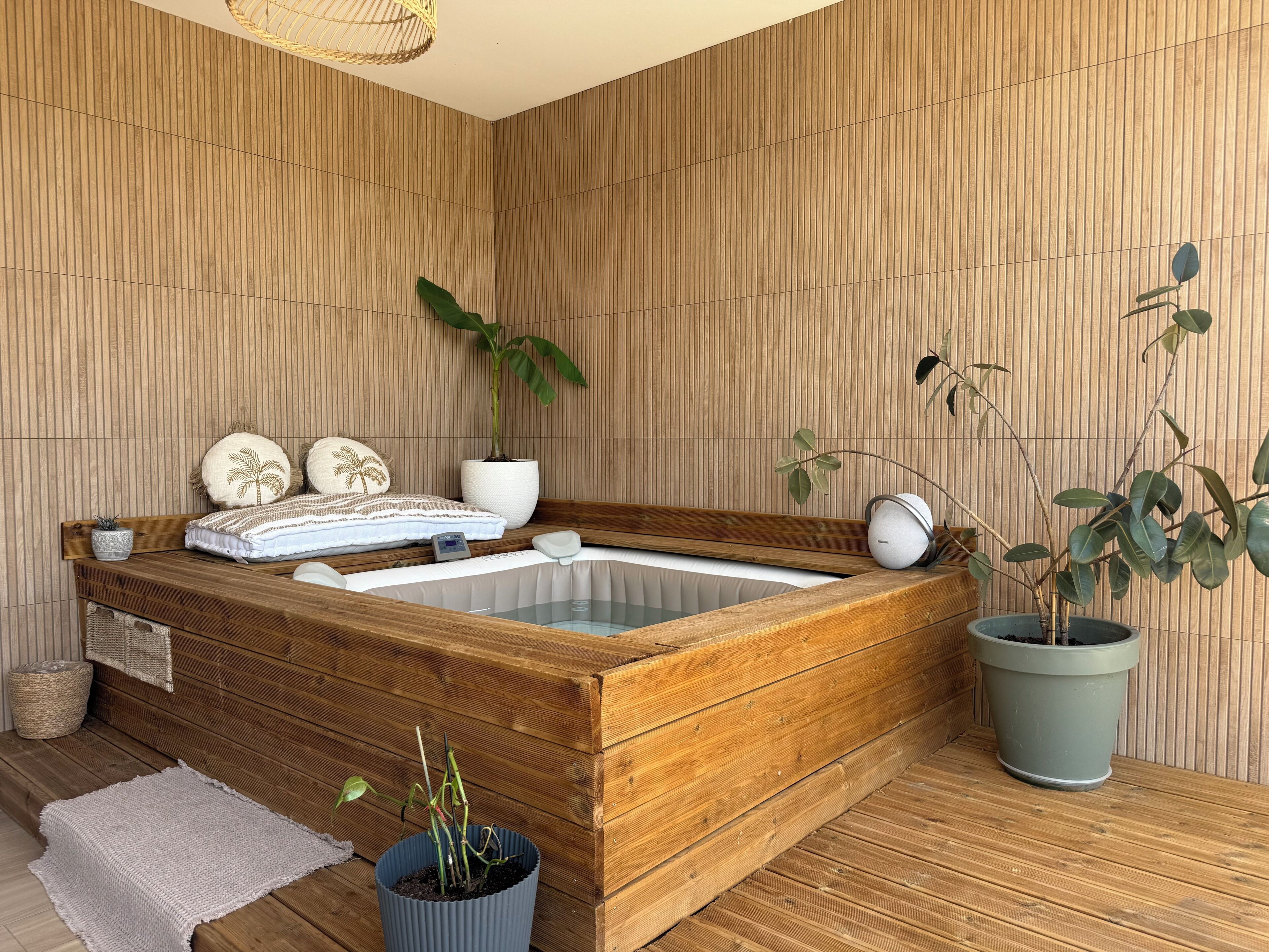 Een houten kuip voor een wellnessmassage met een grote potplant, een klein tafeltje met een plant erop, en een geweven mand.