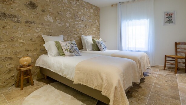 Ein Schlafzimmer mit zwei Betten, einem hölzernen Stuhl, einem kleinen Hocker und einer Wand mit Steinoptik.