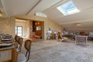 Un loft espacioso con techo alto, paredes de piedra, una zona de comedor con sillas y mesa dispuesta para una comida, un área de estar con sofás y una mesa de centro, y un tragaluz.