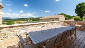Eine Holzterrasse mit Tisch und Stühlen, mit Blick auf eine Steinmauer und eine Hügellandschaft mit Bäumen.