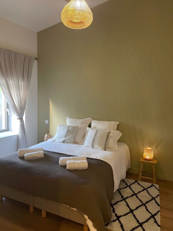 A neatly made bed with white linens and pillows, a dark brown throw blanket, and two folded towels on the bed. There is a small wooden nightstand with a lit lamp beside the bed.