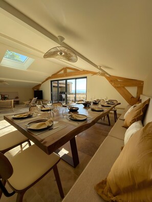 A dining area with a long table set for a meal, a sofa with cushions, and a view through a glass door to the outside.