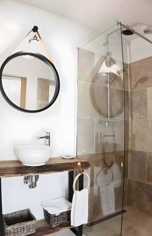 Une salle de bain moderne avec un miroir rond, un lavabo blanc, un plan de toilette en bois, des paniers en osier et une douche Ă parois de verre.