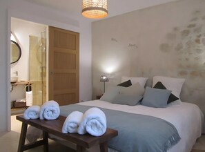 Une chambre Ă coucher comprenant un lit, des oreillers, une lampe de chevet, un banc en bois avec des serviettes, une salle de bain avec un lavabo et un miroir.