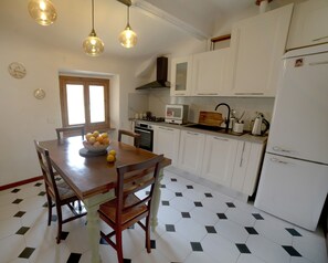 Una cocina con armarios blancos, una mesa de comedor con sillas, un cuenco de fruta y un refrigerador.