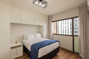 A bedroom with a bed, a nightstand, a window with curtains, and a view of buildings outside. - LTL - L'Etoile Residence by Anora Spaces (Barueri)