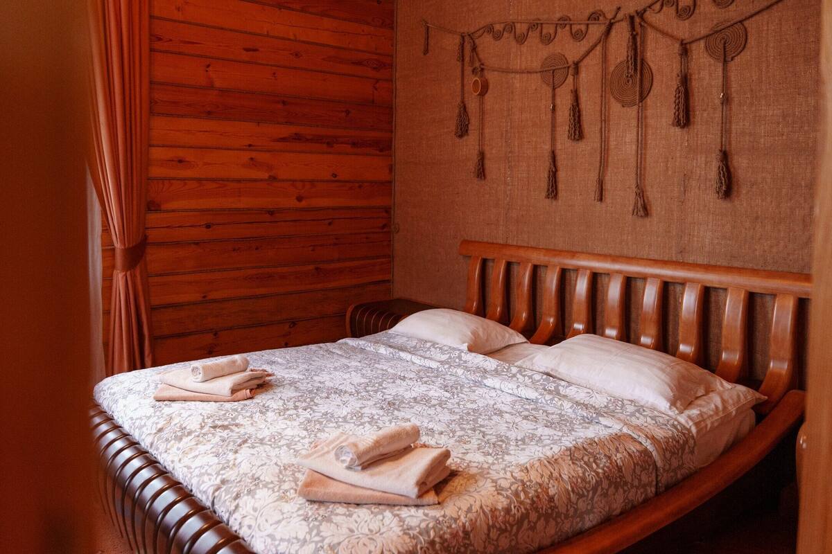 A wooden bed with a patterned bedspread and pillows, a headboard, and a wall with a hanging decoration.