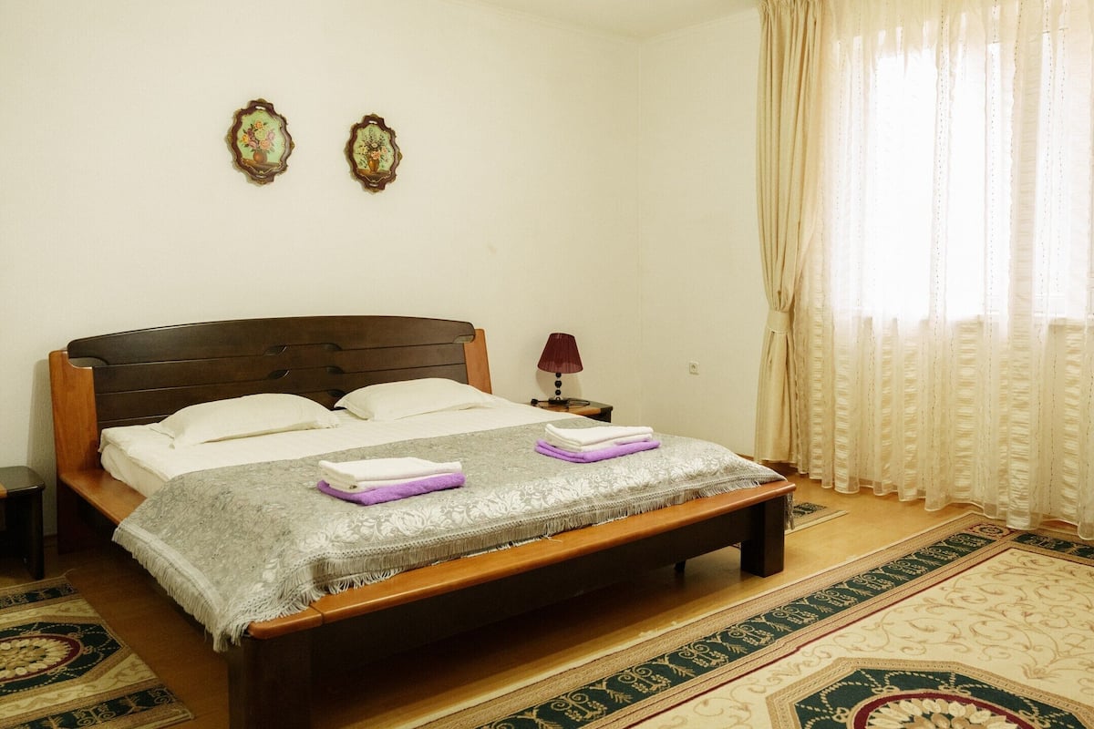 A neatly made bed with a headboard, a bedside table with a lamp, and a decorative rug.