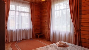 Una habitación de cabaña de madera con dos ventanas grandes, un pequeño taburete de madera y una cama con colcha estampada.
